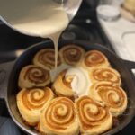 Pouring lemon cream cheese icing onto freshly baked sourdough cinnamon rolls in a cast iron skillet