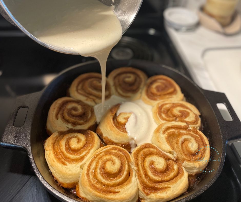 Pouring lemon cream cheese icing onto freshly baked sourdough cinnamon rolls in a cast iron skillet